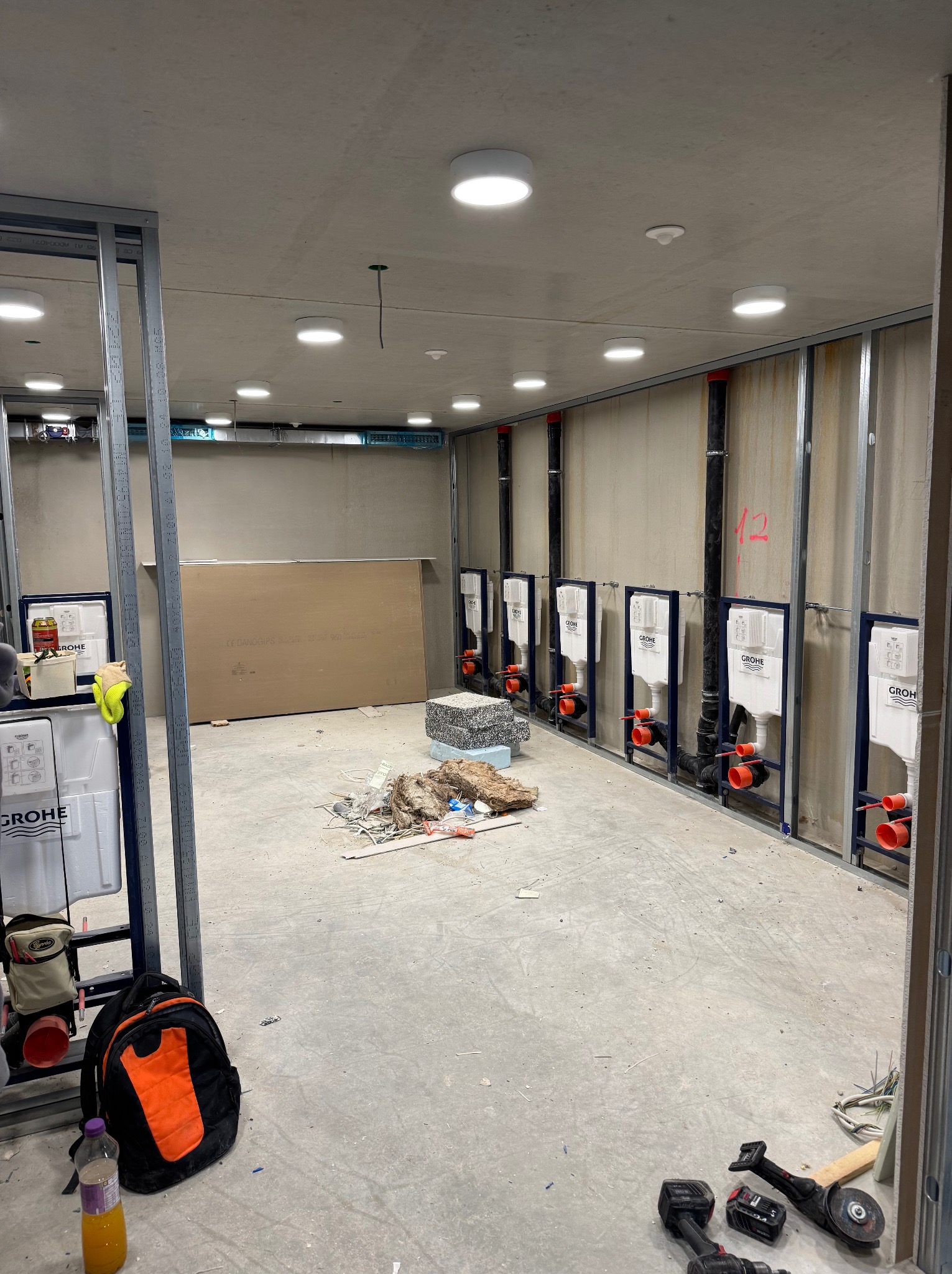Wnętrze w trakcie instalacji podtynkowych stelaży WC Grohe, widoczne rury i narzędzia. Oświetlenie sufitowe w surowym pomieszczeniu z metalowymi konstrukcjami.