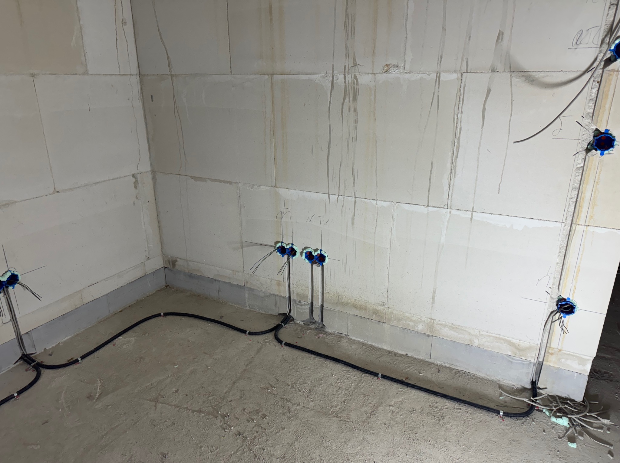 Surowy stan: czarne kable w peszlu prowadzone wzdłuż ścian, niebieskie puszki podtynkowe wystają ze ścian w pomieszczeniu w trakcie prac instalacyjnych.