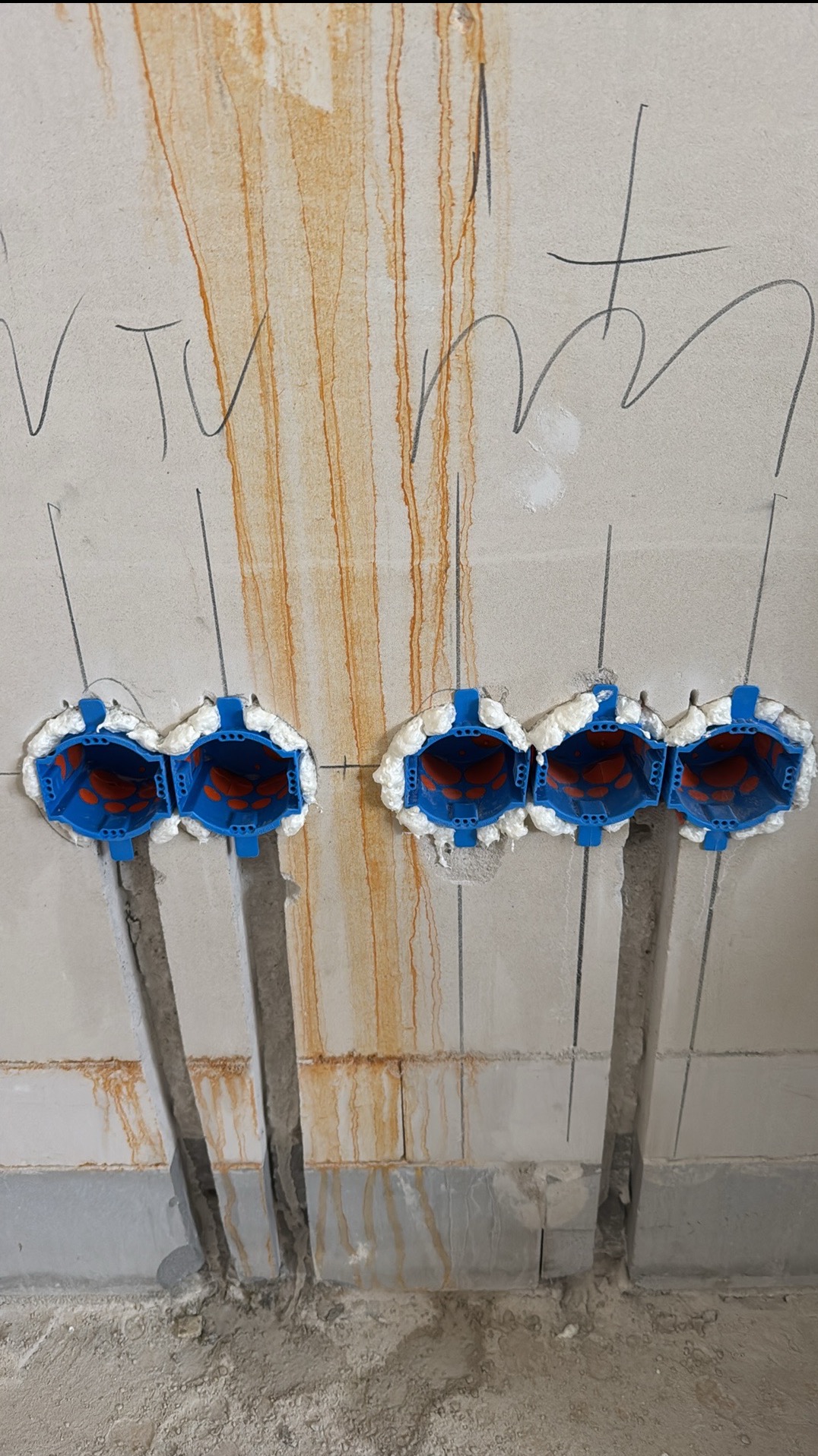 Ściana z wyciętymi otworami pod puszki elektryczne, zabezpieczone pianką montażową. Widoczne pionowe bruzdy pod kable. Ślady po zalaniu na ścianie.