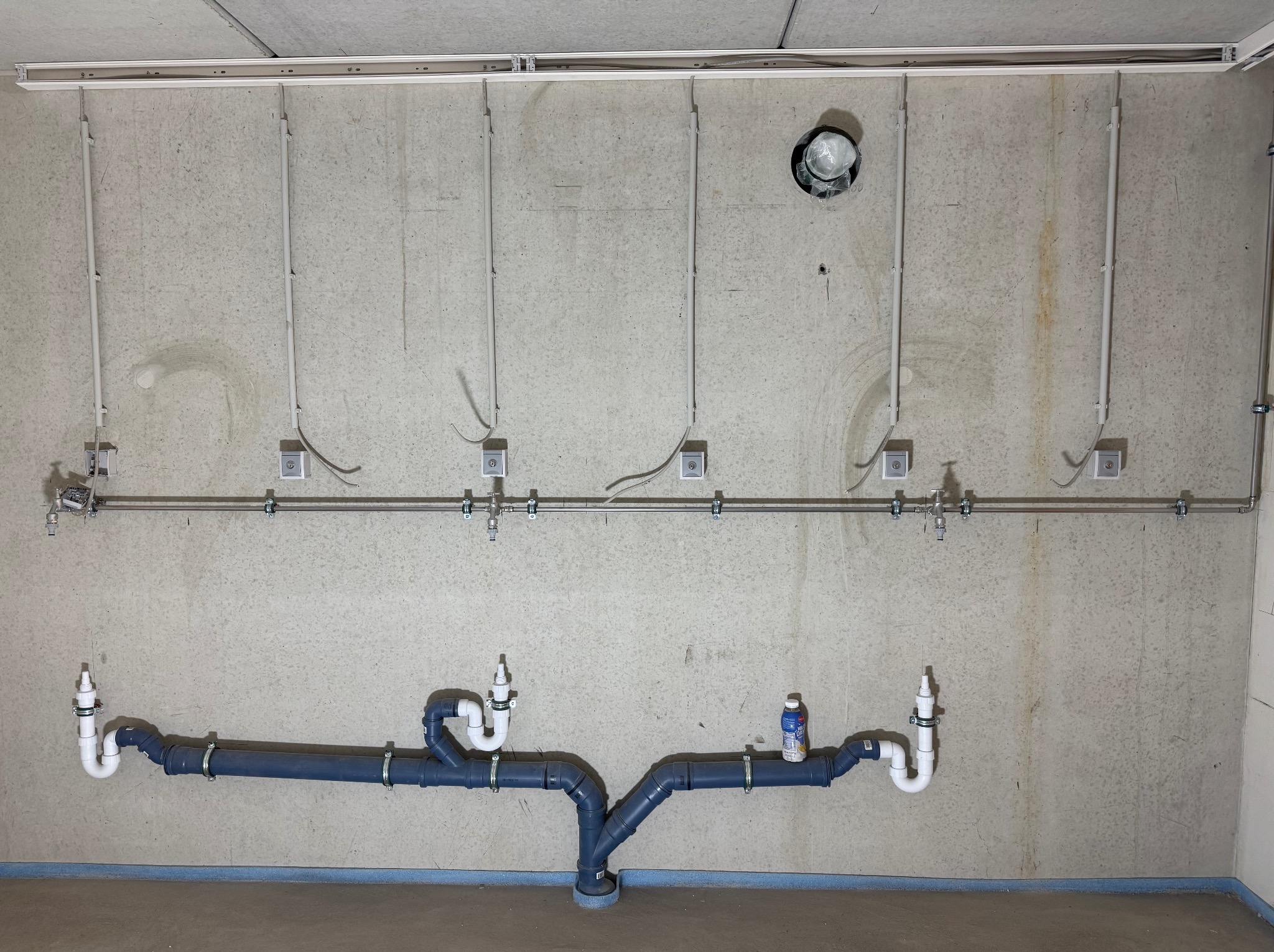 Szara ściana z widocznymi instalacjami: rury kanalizacyjne z syfonami i metalowa listwa z przewodami elektrycznymi. Surowy stan wykończenia wnętrza.