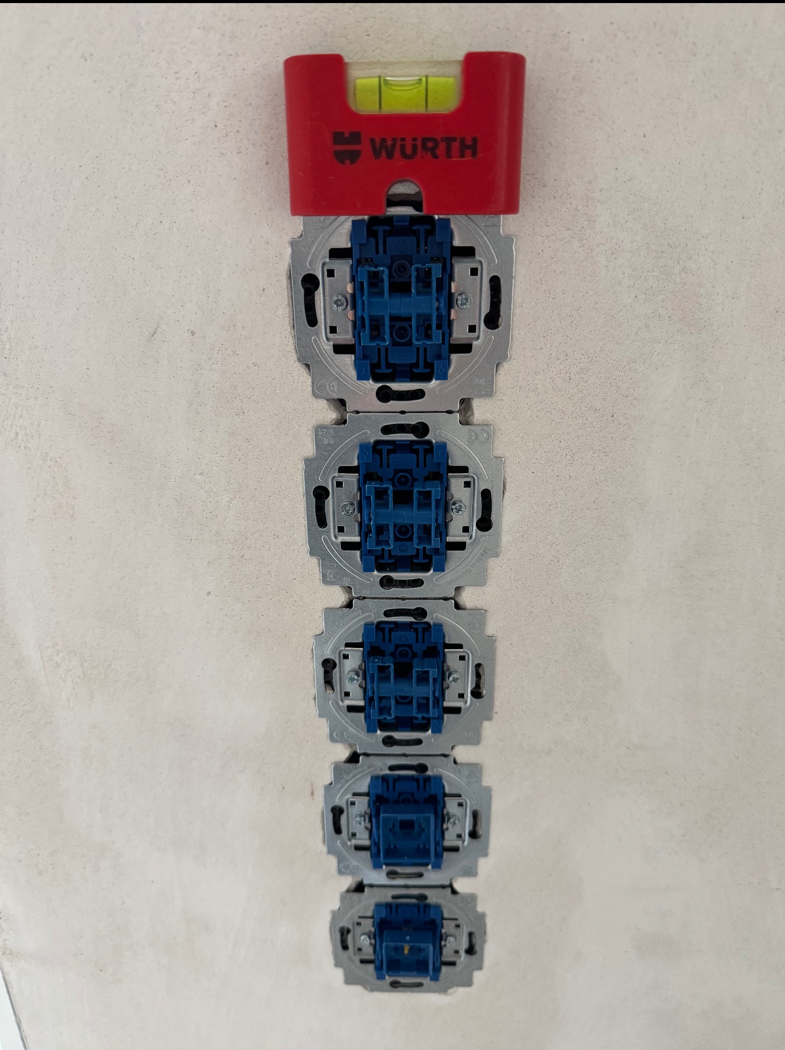 Rząd gniazdek elektrycznych z niebieskimi wkładami, wyrównanych pionowo za pomocą poziomicy marki Wurth na tle jasnej ściany. Widok z bliska na etapie montażu.