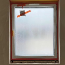 Bohdan Shulzhenko - Białe okno zabezpieczone folią i taśmą malarską w trakcie prac wykończeniowych. Widoczne gniazdka elektryczne pod oknem i fragment instalacji na suficie.