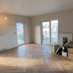 Bohdan Shulzhenko - Jasne, przestronne wnętrze po remoncie z parkietem ułożonym w jodełkę. Widoczne okna balkonowe i grzejniki. Część materiałów budowlanych w kartonach w rogu pokoju.