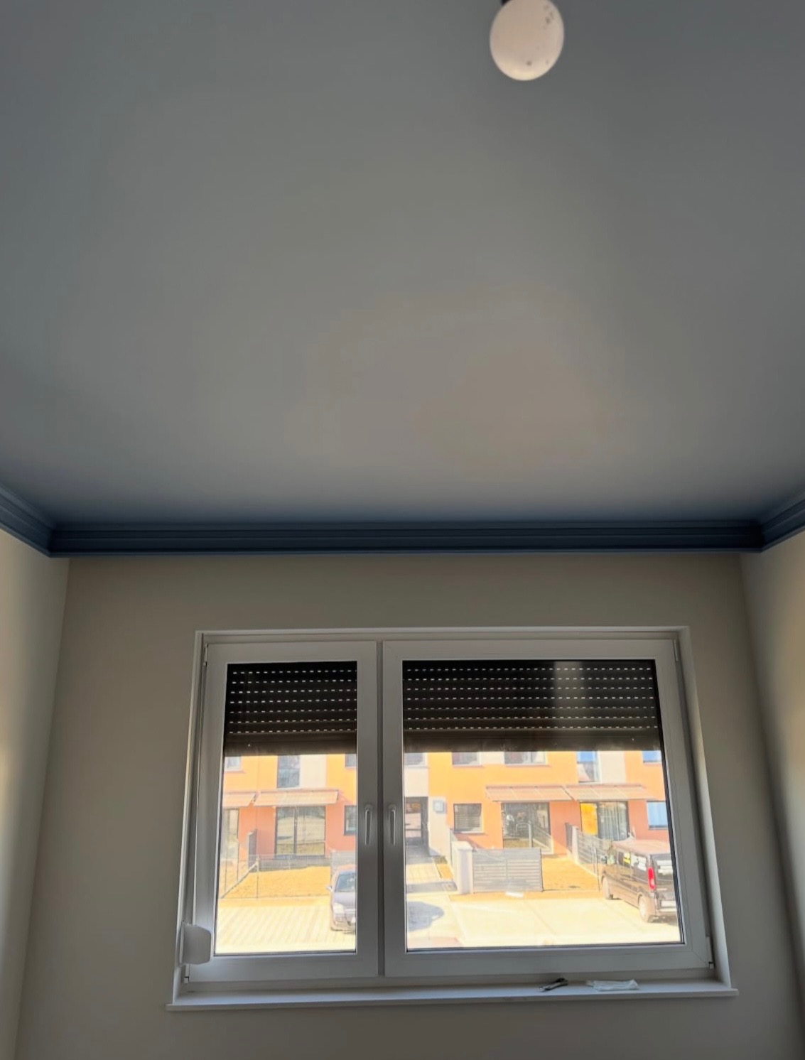 Pomieszczenie po malowaniu: białe ściany, niebieski sufit z listwą, okno z roletami. Widok na budynek z zewnątrz. Nowoczesne wykończenie wnętrza.
