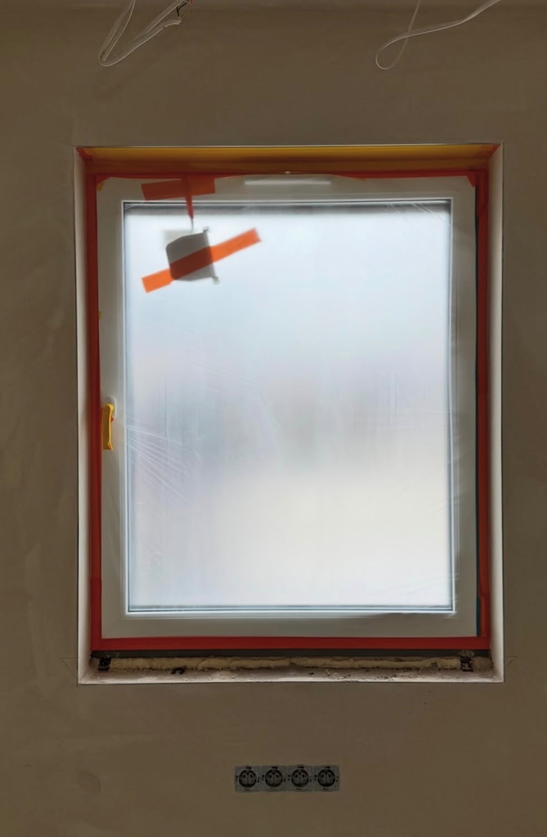Białe okno zabezpieczone folią i taśmą malarską w trakcie prac wykończeniowych. Widoczne gniazdka elektryczne pod oknem i fragment instalacji na suficie.