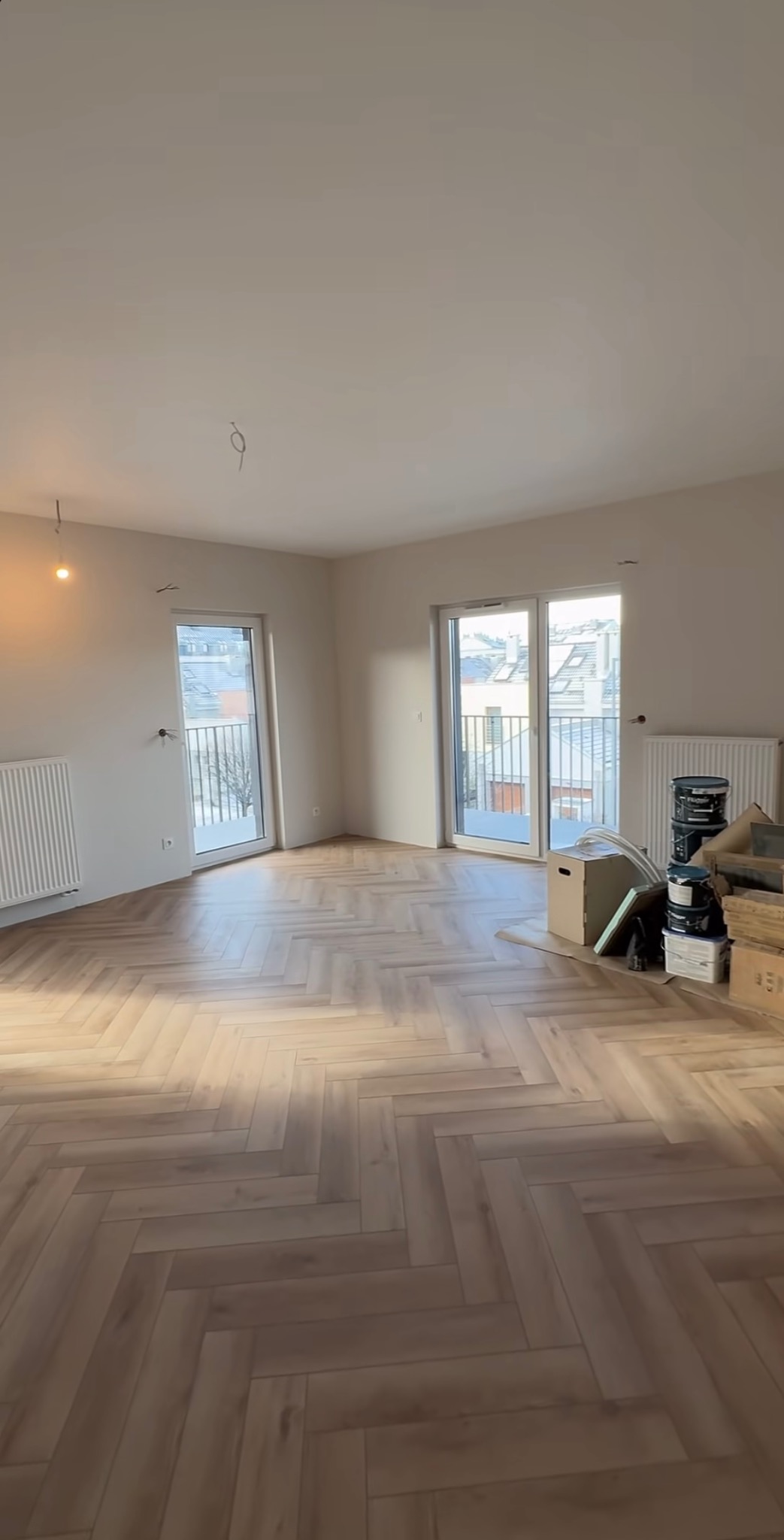 Jasne, przestronne wnętrze po remoncie z parkietem ułożonym w jodełkę. Widoczne okna balkonowe i grzejniki. Część materiałów budowlanych w kartonach w rogu pokoju.