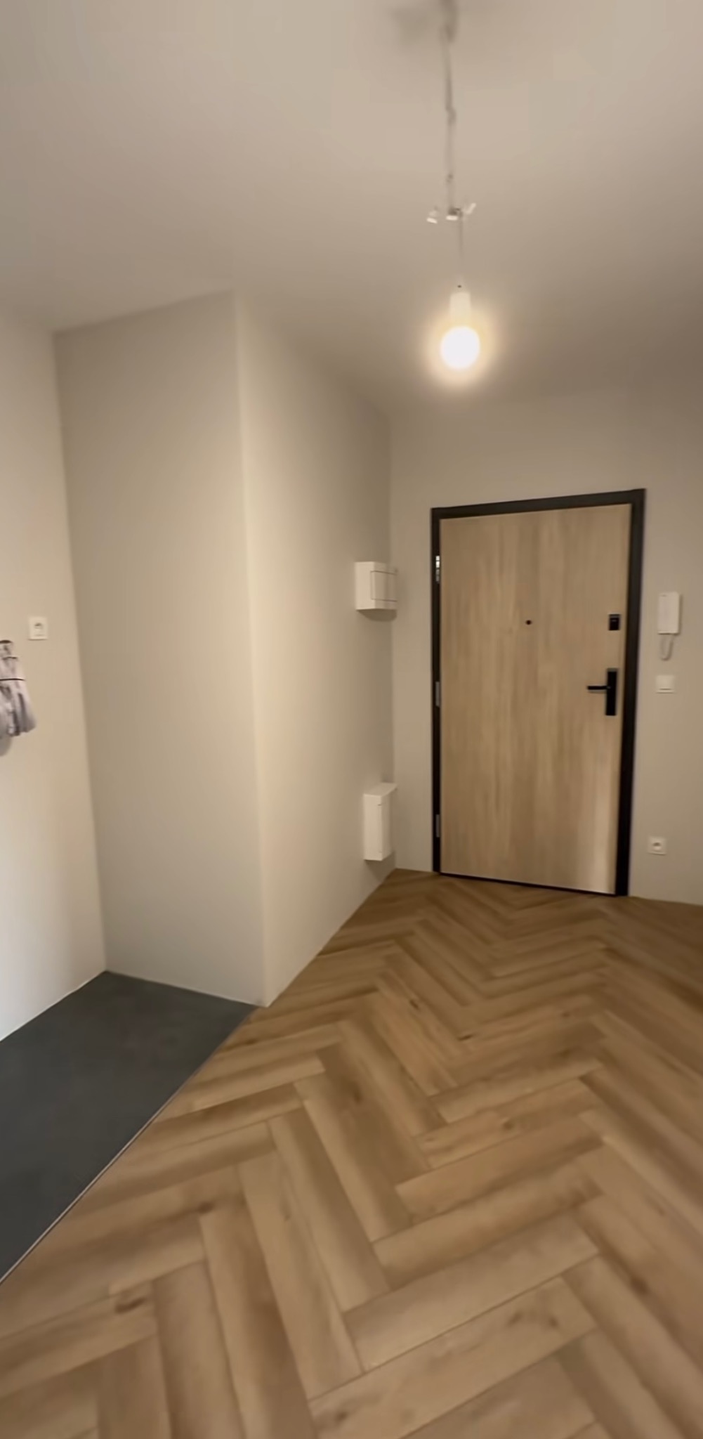 Jasne panele ułożone w jodełkę w przedpokoju, widok na drzwi wejściowe i fragment szarej podłogi. Minimalistyczne wnętrze z oświetleniem punktowym.