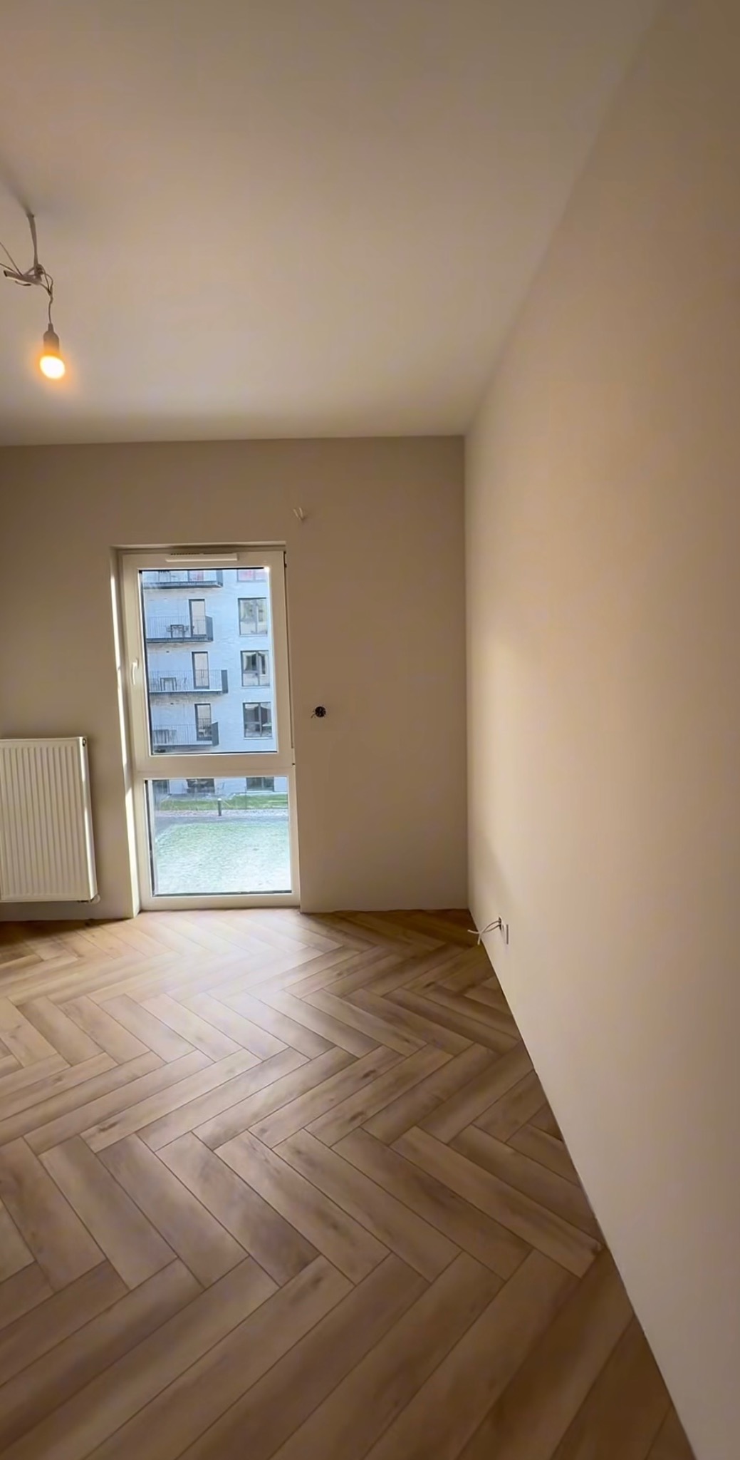 Jasne wnętrze po remoncie: parkiet ułożony w jodełkę, świeżo pomalowane ściany, okno z widokiem na budynek. Widoczne gniazdka elektryczne i grzejnik.
