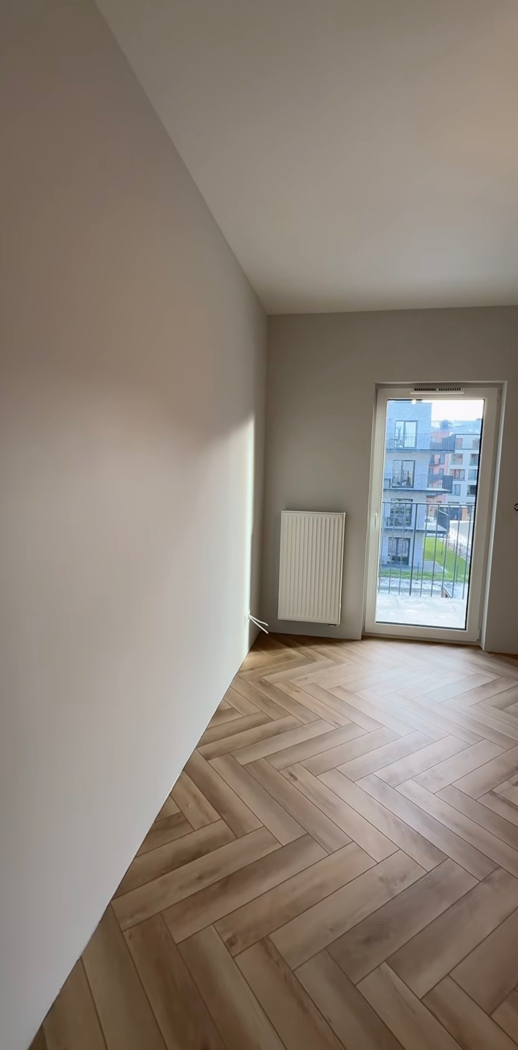 Jasne wnętrze po remoncie: świeżo pomalowane ściany w odcieniach beżu, parkiet ułożony w jodełkę, biały grzejnik i okno z widokiem na budynek mieszkalny.