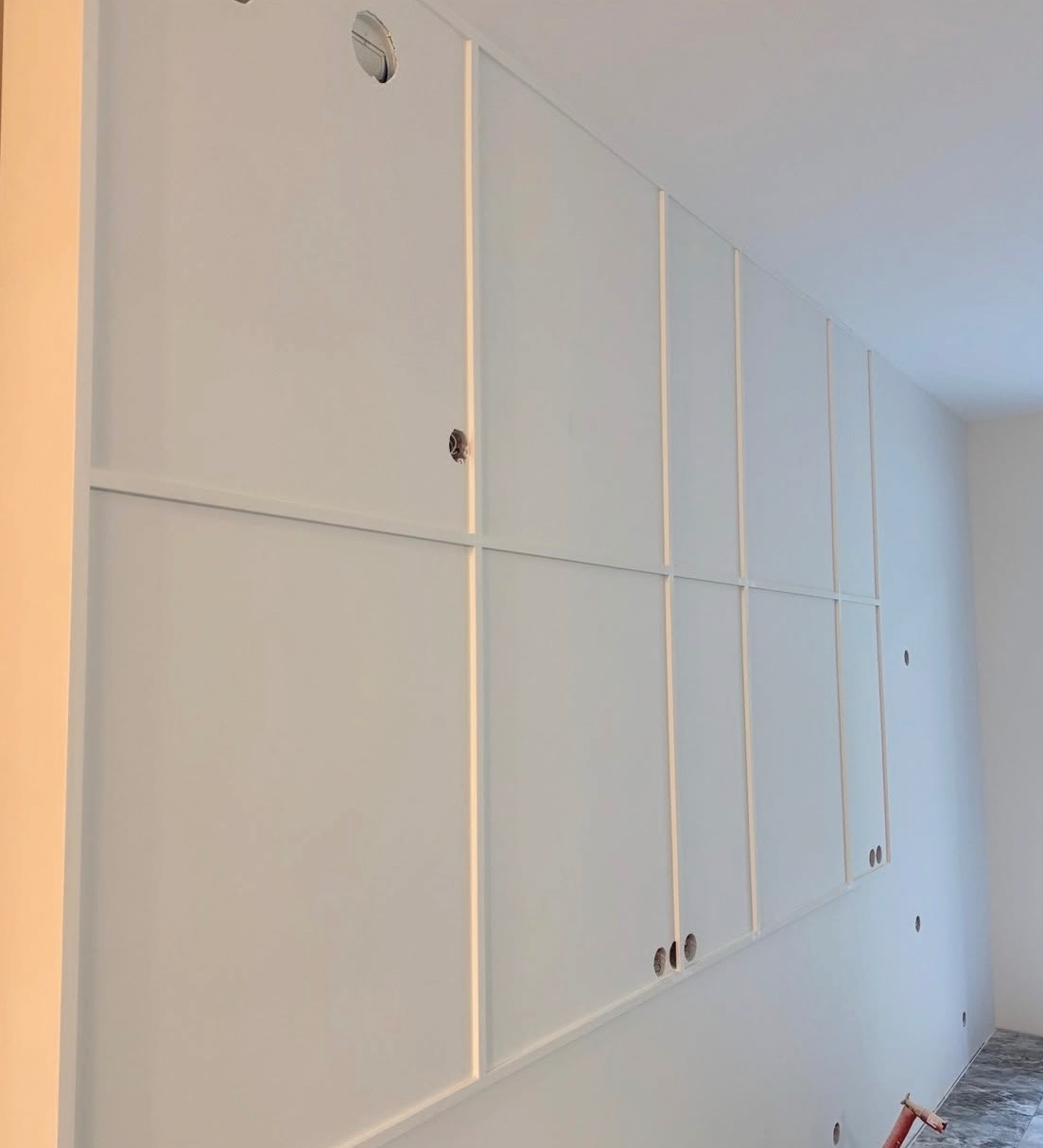 Biała ściana z dekoracyjnymi listwami i otworami na gniazdka, w trakcie wykańczania wnętrza. Widoczny fragment podłogi i narzędzia.