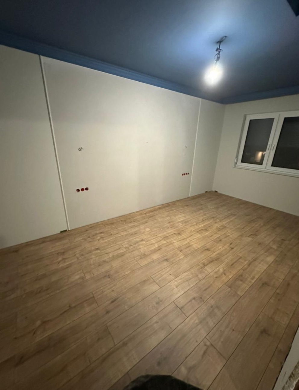 Jasne panele podłogowe w pokoju z białymi ścianami i niebieskim sufitem. Widoczne gniazdka elektryczne i okno z białymi ramami. Oświetlenie sufitowe.