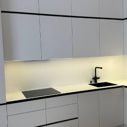 Profi Meble Oleksii Sainchyn - Minimalistyczna kuchnia w bieli z czarnymi akcentami: zabudowa z szafkami, oświetlenie LED pod szafkami, czarna armatura i płyta indukcyjna. Nowoczesny design.
