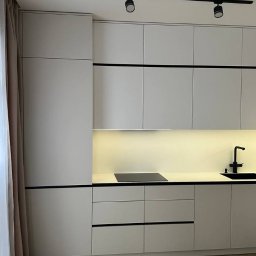Profi Meble Oleksii Sainchyn - Minimalistyczna kuchnia z jasnymi frontami, czarnymi akcentami i oświetleniem LED. Widoczny fragment stołu i krzesła. Nowoczesny design.