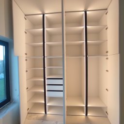 Profi Meble Oleksii Sainchyn - Otwarta, biała szafa wnękowa z oświetleniem LED, widoczne półki i szuflady. Minimalistyczny design, nowoczesne wykończenie, surowe wnętrze w tle.