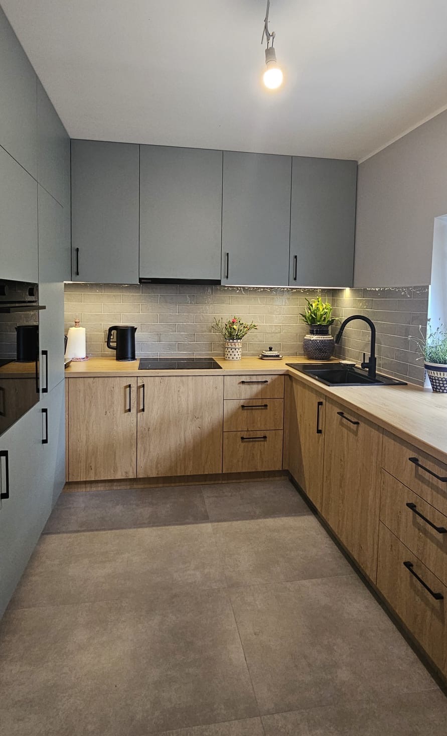 Nowoczesna kuchnia z szarymi szafkami i drewnianymi blatami. Czarny zlew i armatura, płytki imitujące cegłę. Oświetlenie podszafkowe i dekoracje roślinne.