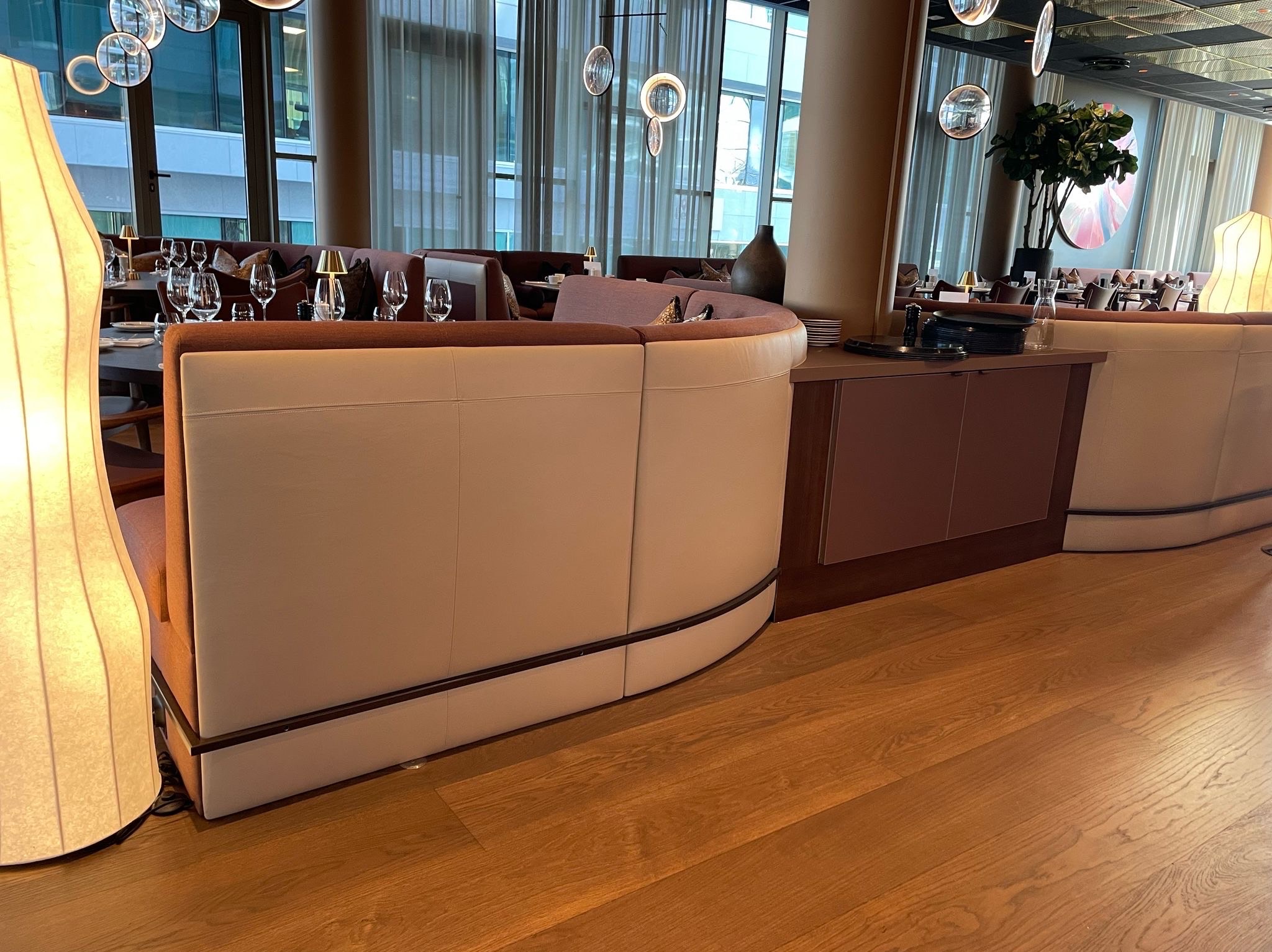 Eleganckie, półokrągłe sofy z jasną tapicerką i ciemnym wykończeniem w restauracji. Widoczne nakryte stoły, lampy i drewniana podłoga. Nowoczesny design wnętrza.