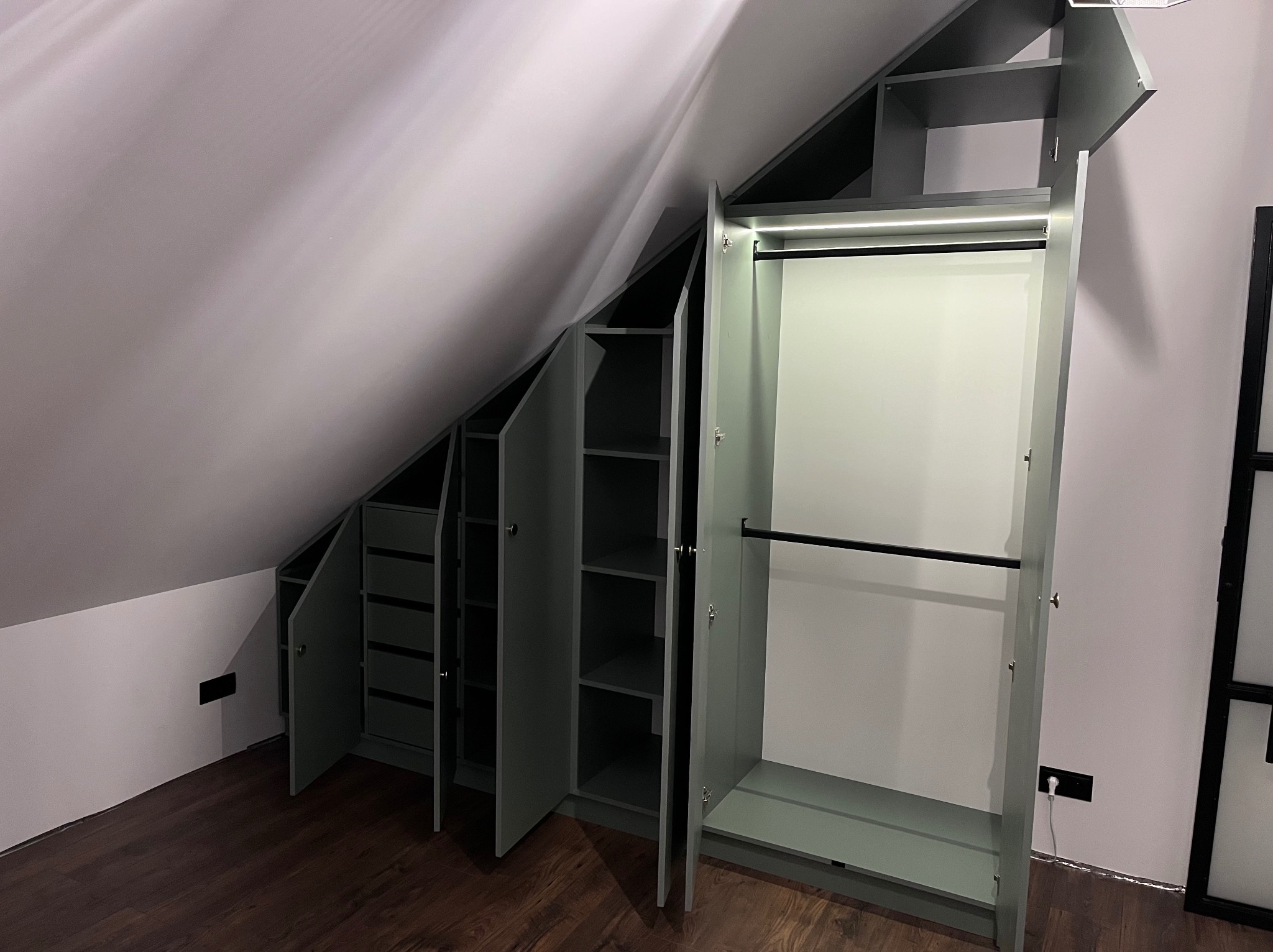 Szara szafa wnękowa na poddaszu ze skośnym sufitem, z otwartymi drzwiami ukazującymi półki i drążek na ubrania, oświetlona listwą LED.