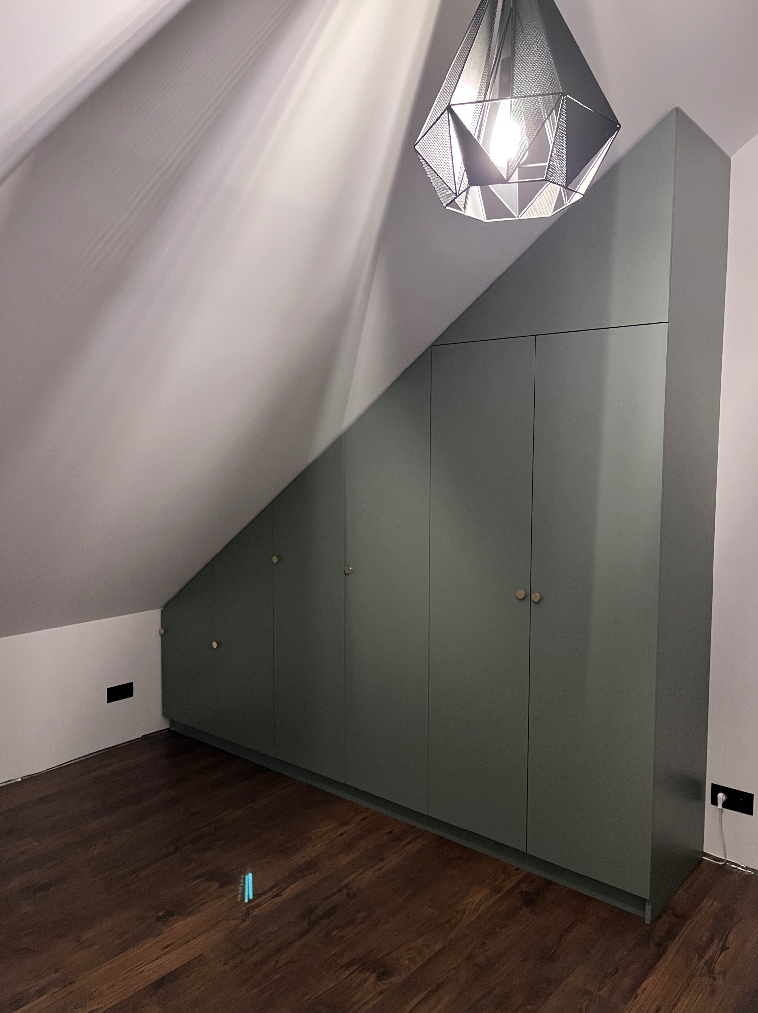 Szafa wnękowa w kolorze oliwkowym, dopasowana do skosu poddasza, z okrągłymi uchwytami. Na suficie designerska lampa. Podłoga z paneli w odcieniach brązu.