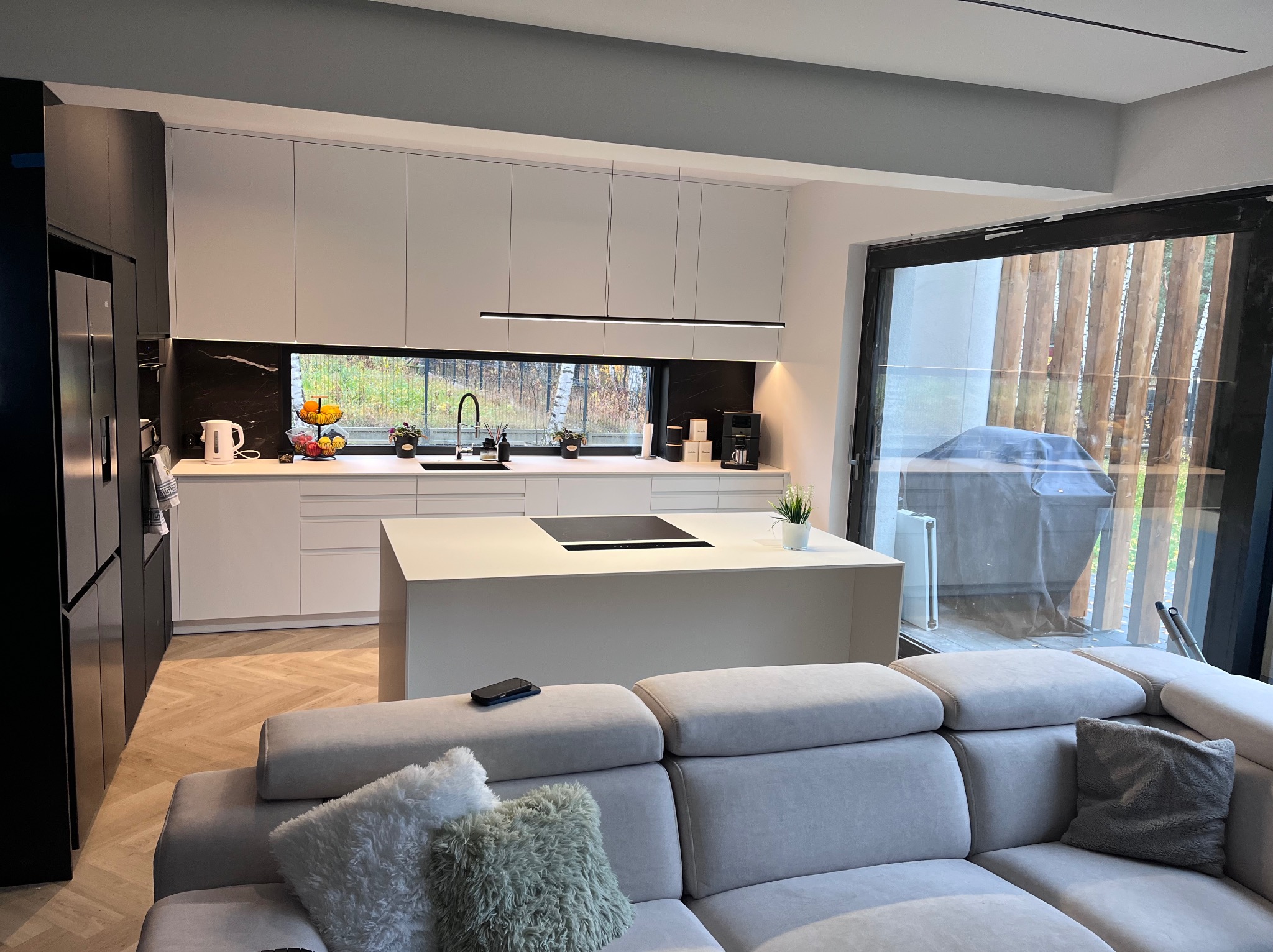 Nowoczesna kuchnia z wyspą i białymi szafkami, czarne akcenty, widok z salonu. Parkiet w jodełkę, duże okno z widokiem na zewnątrz.