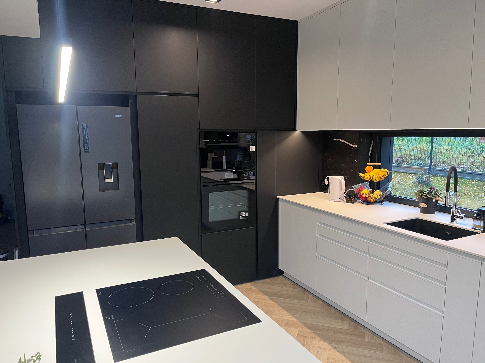 Nowoczesna kuchnia: matowe, czarne szafki kontrastują z białym blatem i frontami. Widoczny sprzęt AGD, okno z widokiem na zieleń i drewniana podłoga w jodełkę.