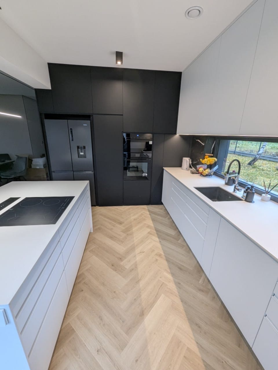 Nowoczesna kuchnia z białymi szafkami i wyspą, czarne szafki wiszące, parkiet w jodełkę. Minimalistyczny design, okno z widokiem na zieleń.