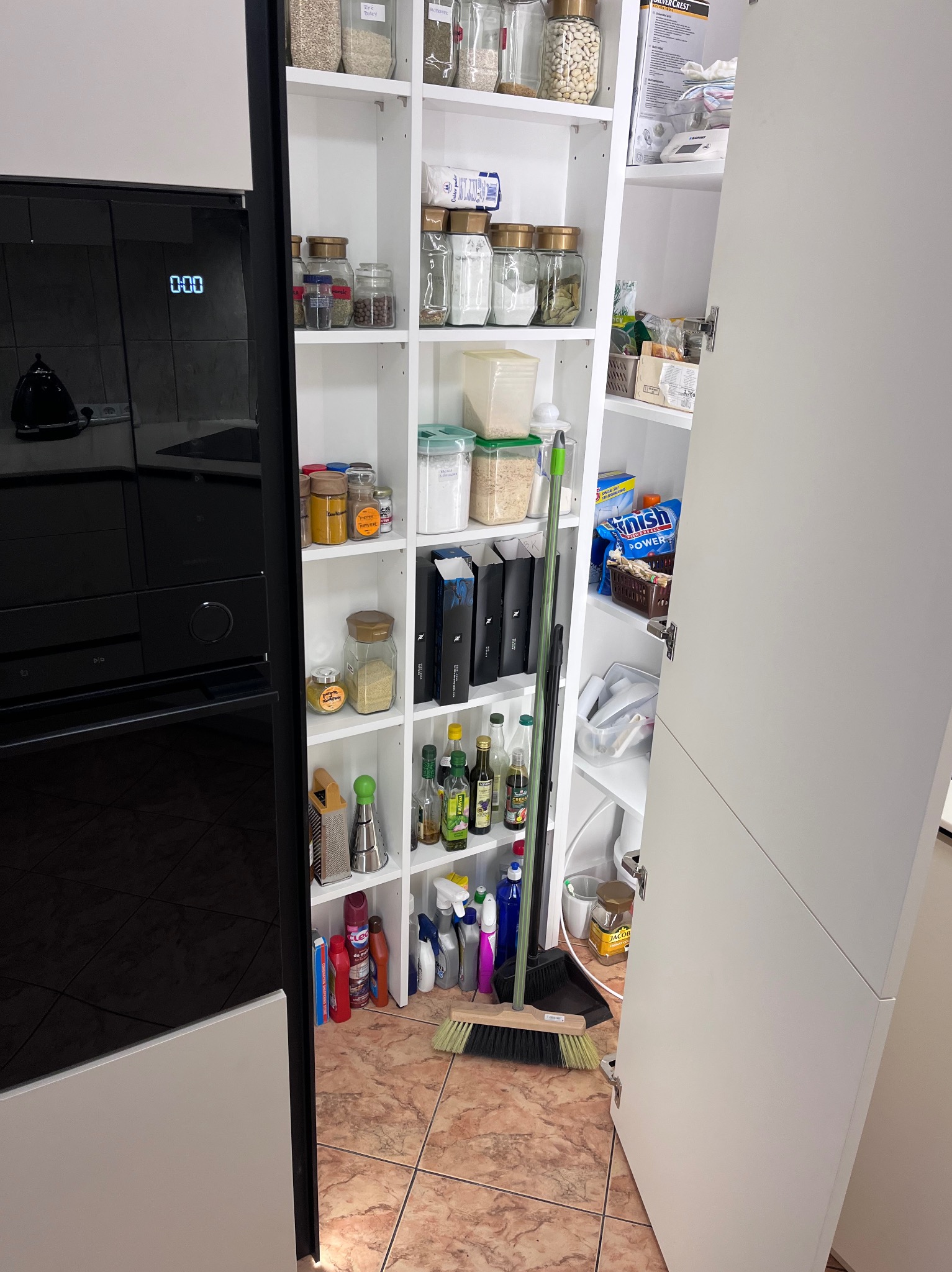 Biała spiżarnia z półkami pełnymi słoików, pojemników i środków czystości, widoczna obok czarnego piekarnika w nowoczesnej kuchni.