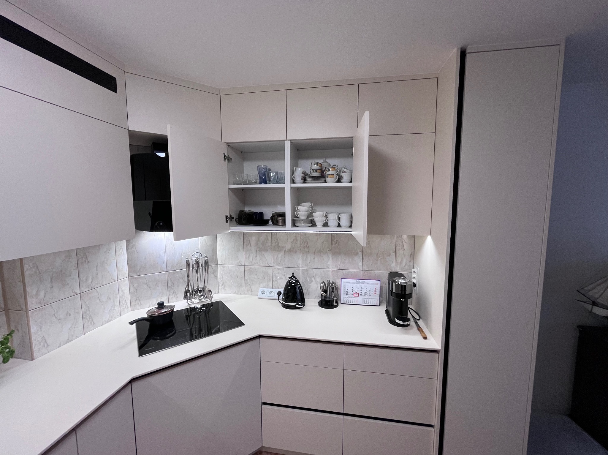 Jasna kuchnia z otwartą szafką, w której widoczne są naczynia. Na blacie czajnik, ekspres do kawy i kalendarz. Minimalistyczny design, marmurowe płytki na ścianie.