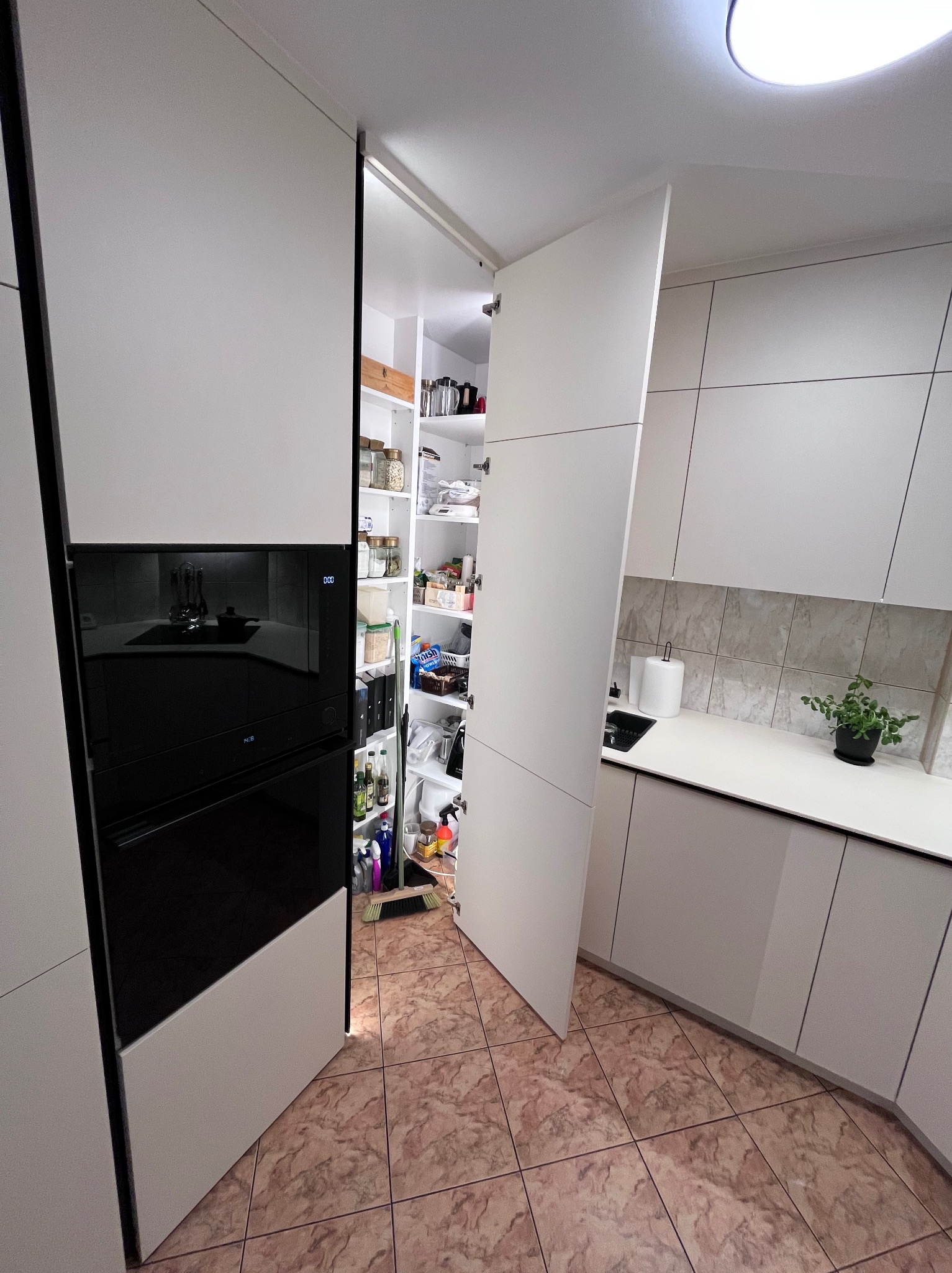 Nowoczesna kuchnia z białymi frontami i ukrytą spiżarnią za przesuwnymi drzwiami. Widoczny piekarnik w zabudowie i blat z rośliną doniczkową.