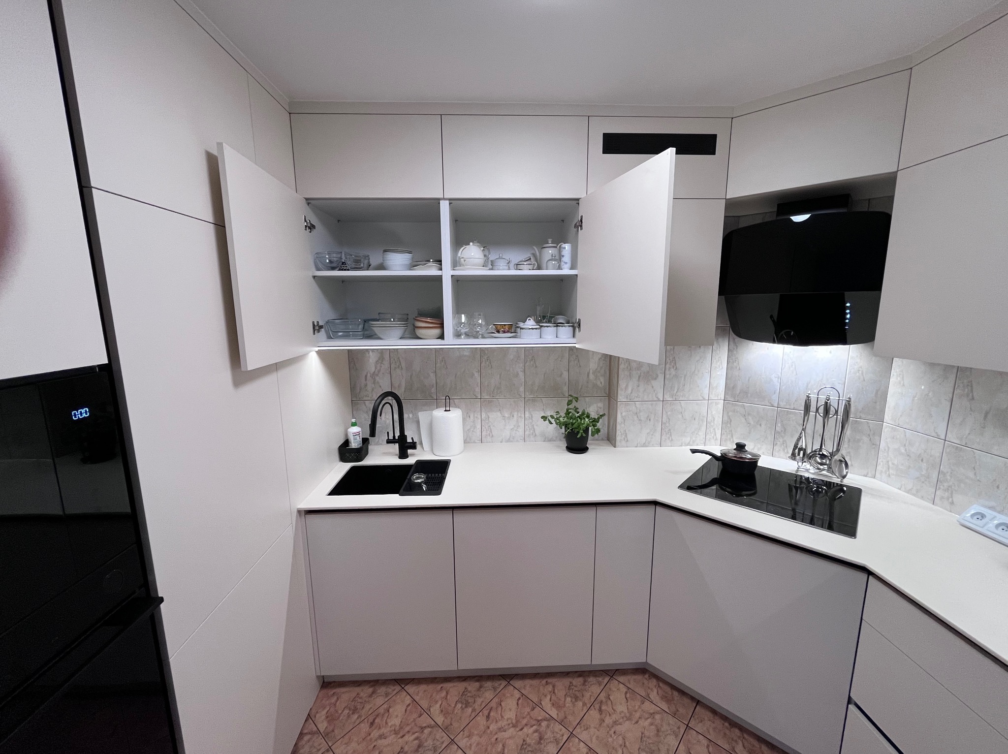 Biała, nowoczesna kuchnia z czarnym zlewem i płytą grzewczą. Otwarta szafka ukazuje zastawę. Minimalistyczny design, jasne wnętrze, płytki w tle.