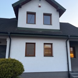 Włodzimierz Koza - Dwukondygnacyjny dom z białymi ścianami, brązowymi ramami okiennymi i ciemnym dachem. Widoczne okna z żaluzjami i krzew przed budynkiem. Ujęcie z dołu.