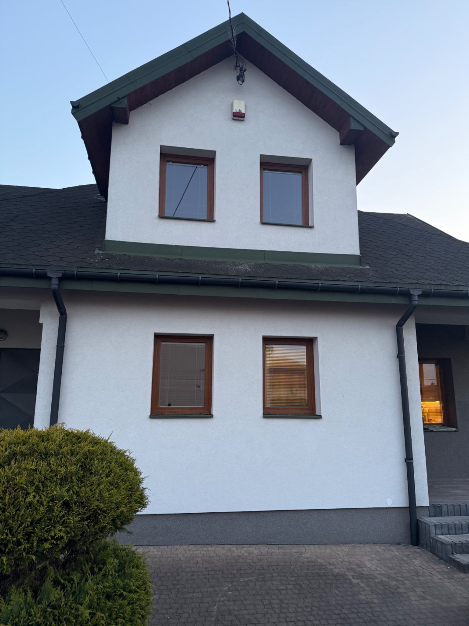 Dwukondygnacyjny dom z białymi ścianami, brązowymi ramami okiennymi i ciemnym dachem. Widoczne okna z żaluzjami i krzew przed budynkiem. Ujęcie z dołu.