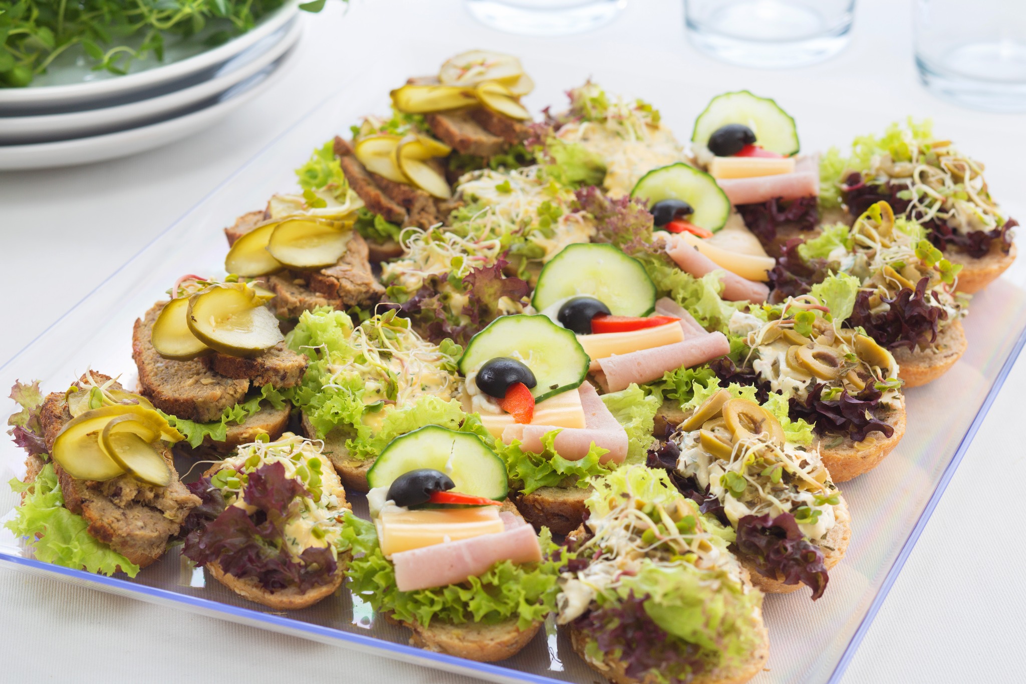 Dekoracyjne kanapeczki na przezroczystej tacy, z różnymi dodatkami: ogórki, szynka, ser, oliwki, sałata i kiełki. Idealne na catering firmowy lub imprezę okolicznościową.