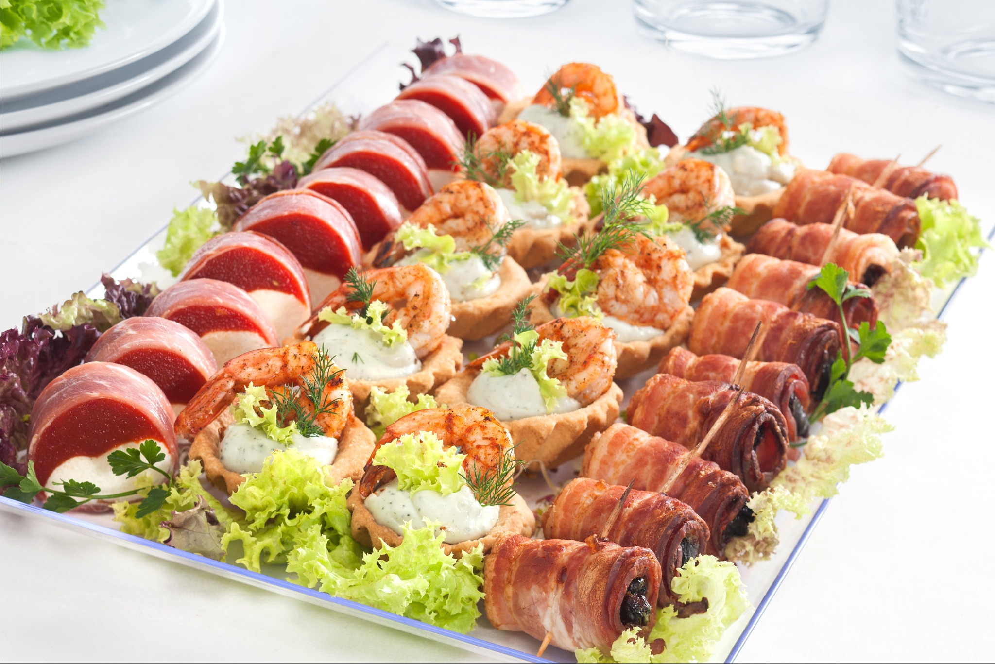 Elegancka taca z wykwintnymi przekąskami: roladki z szynki, tartaletki z krewetkami i bekonowe ruloniki z suszonymi śliwkami, udekorowane świeżą zieleniną.