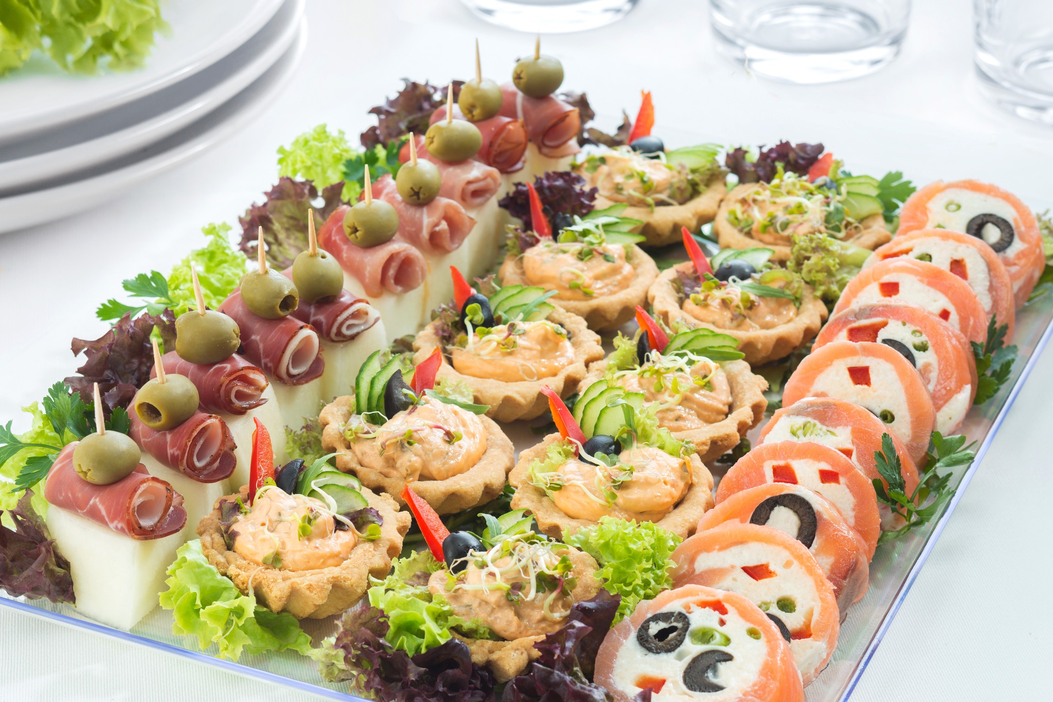 Elegancka taca z różnorodnymi przekąskami: tartaletki z pastą, roladki z łososia i koreczki z szynki parmeńskiej i oliwek, na białym tle. Idealne na przyjęcie.