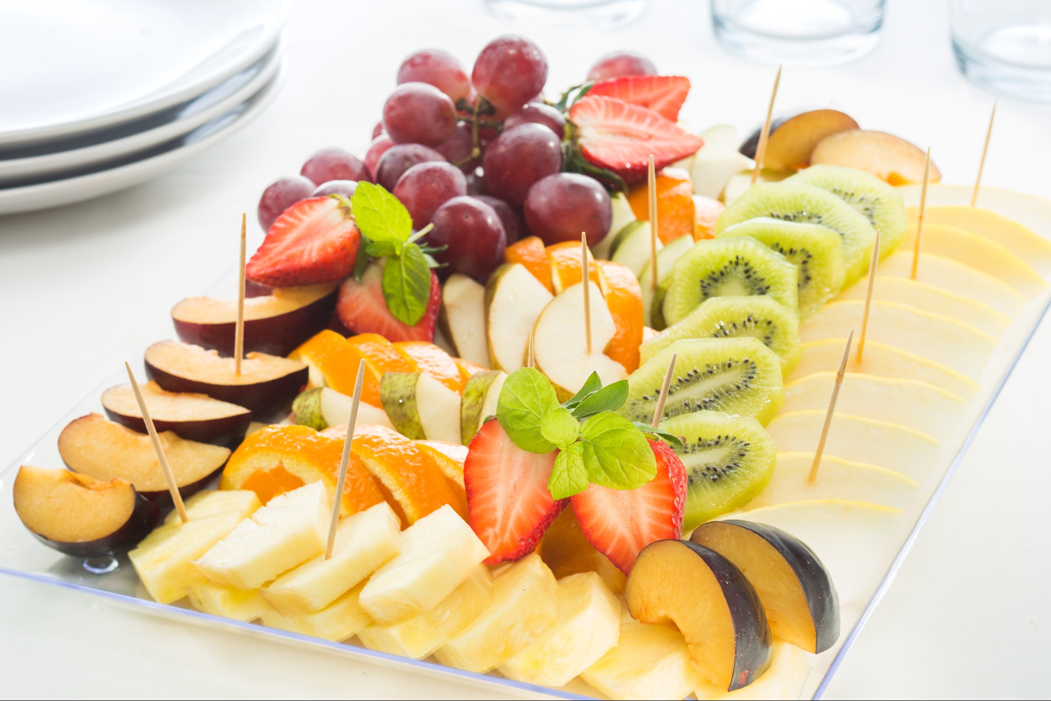 Elegancka taca z owocami: winogrona, truskawki, kiwi, śliwki, ananas i pomarańcze, udekorowane listkami mięty, na białym tle. Idealne na przyjęcie.