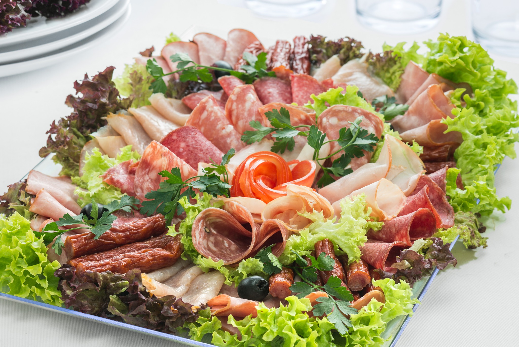 Dekoracyjna taca z wędlinami, salami i sałatą lodową. Kompozycja z roladek szynki, kiełbasy i ziół na białym stole. Elegancki catering na imprezę.