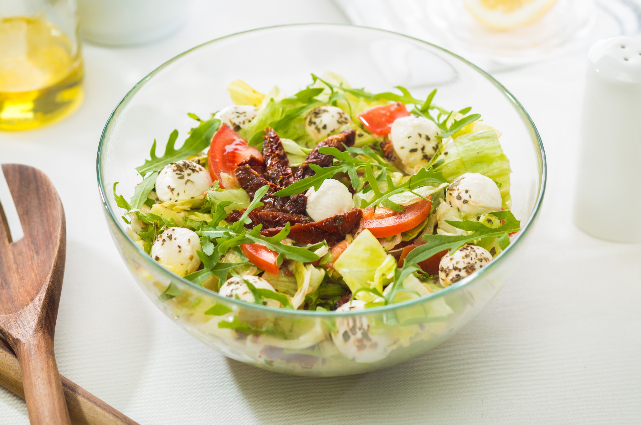 Świeża sałatka w szklanej misce: mozzarella, pomidory, rukola i suszone pomidory na białym tle. Drewniane sztućce do sałatek obok.