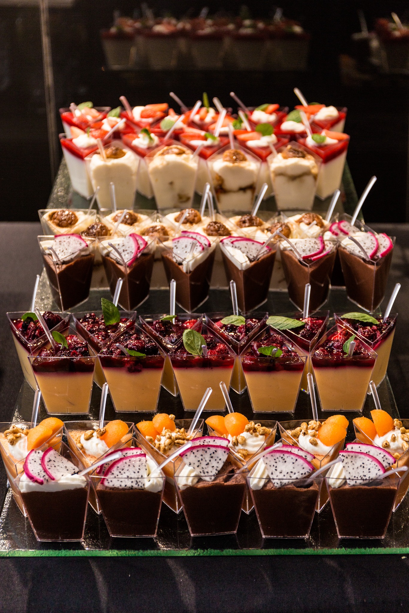 Elegancki bufet deserowy: różnorodne mini desery w przezroczystych naczyniach, udekorowane owocami (truskawki, smoczy owoc, morele) i bitą śmietaną, na szklanych półkach.