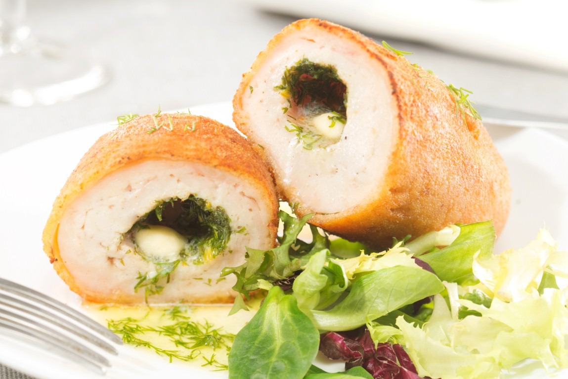 Dwa przekrojone roladki drobiowe z ziołowym farszem i serem, ułożone na białym talerzu z sałatką. Widoczny widelec i delikatne oświetlenie.