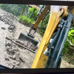 Roboty ziemne 24 - Żółta koparka z łyżką pełną kostki brukowej na piasku, w tle zielona roślinność i fragment budynku. Prace ziemne w toku, przygotowanie podłoża pod nową nawierzchnię.