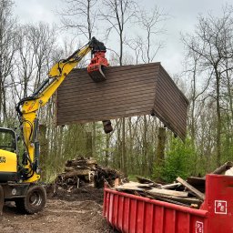 Roboty ziemne 24 - Żółta koparka Wacker Neuson z chwytakiem demontuje drewnianą konstrukcję na tle lasu i czerwonego kontenera na gruz. Widoczne ślady po pracy ziemnej.