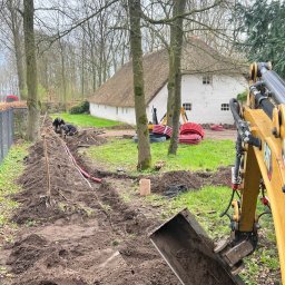 Roboty ziemne 24 - Wykop pod instalacje: żółta koparka w akcji, widoczne rury i pracownicy w tle, dom z strzechą. Prace ziemne w toku, teren przygotowywany pod ułożenie instalacji.