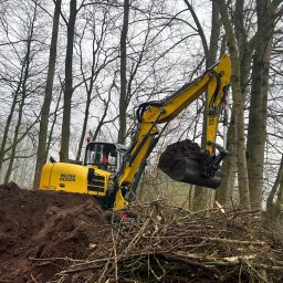Roboty ziemne 24 - Żółta koparka Wacker Neuson w lesie, nabiera ziemię z wykopu obok sterty gałęzi. Widoczne drzewa bez liści i pochmurne niebo w tle. Praca ziemna w toku.