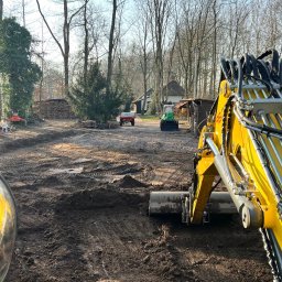 Roboty ziemne 24 - Żółta koparka w trakcie prac ziemnych w Józefowie, widok z kabiny. W tle drzewa i zabudowania, teren przygotowany pod dalsze prace.