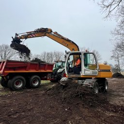 Roboty ziemne 24 - Żółta koparka Liebherr ładuje ziemię z korzeniami do czerwonej przyczepy na tle pola i drzew, widoczny operator w kabinie.