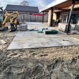 Roboty ziemne 24 - Układanie szarych płyt na tarasie w ogrodzie. Mini koparka CAT i palety z kostką w tle. Częściowo ułożona nawierzchnia, widoczne narzędzia i poziomica.
