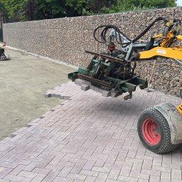 Roboty ziemne 24 - Układanie kostki brukowej maszyną. Żółty podnośnik z zielonym chwytakiem układa kostkę w wzór jodełki. W tle przygotowany teren i mur z kamieni.