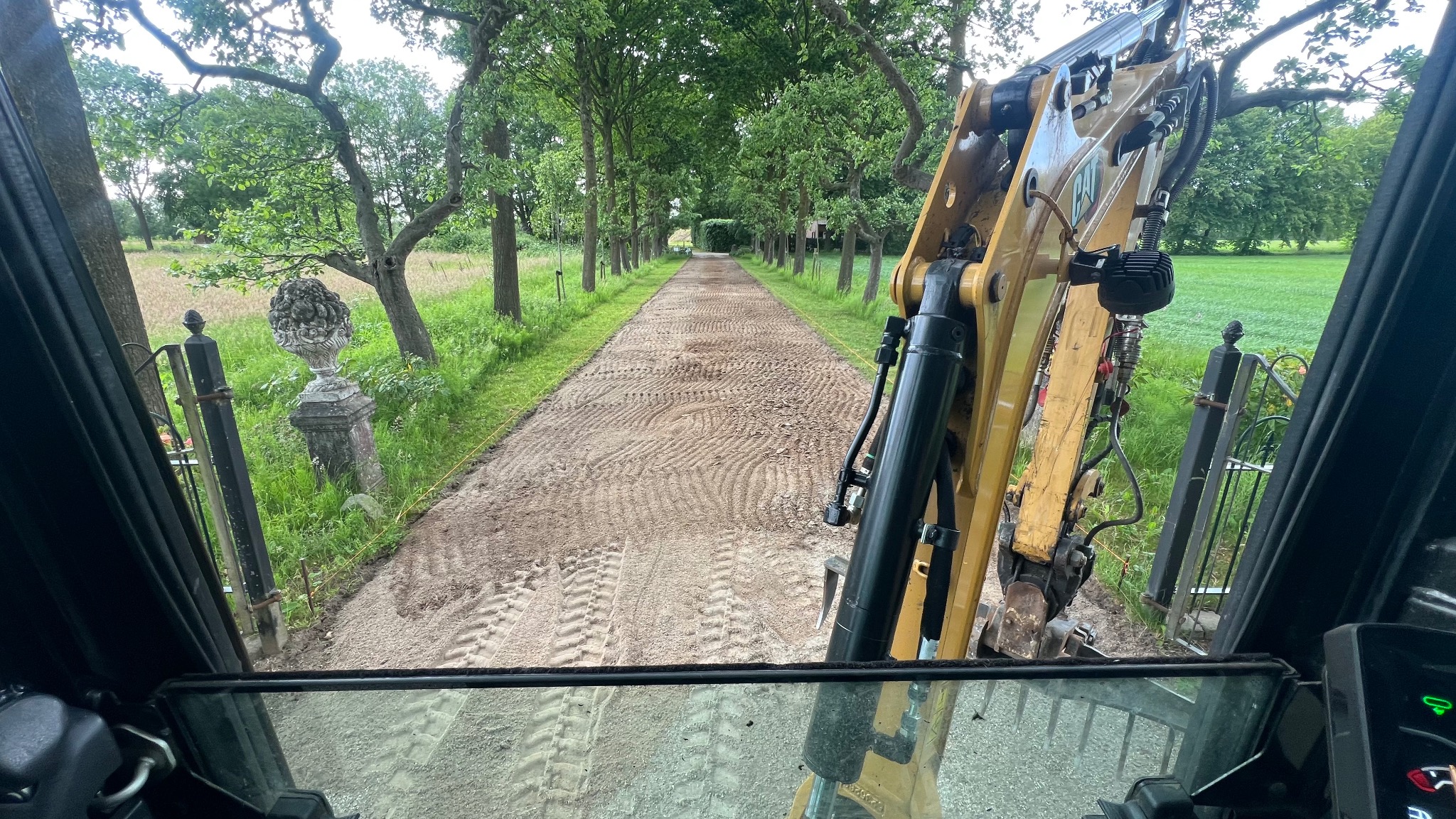 Widok z kabiny koparki na wyrównany, piaszczysty teren alei parkowej z rzędami drzew. W tle brama z ozdobnymi wazonami. Prace ziemne w otoczeniu zieleni.
