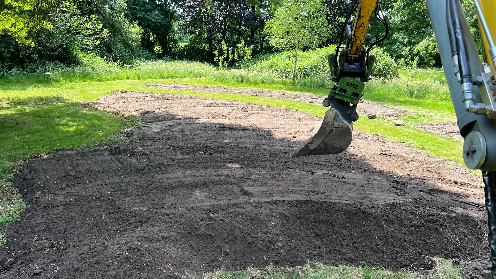 Żółta koparka SMZ w trakcie niwelacji terenu pod przyszły trawnik. Wykopana ziemia tworzy ciemny kontrast z zielenią trawy w tle.