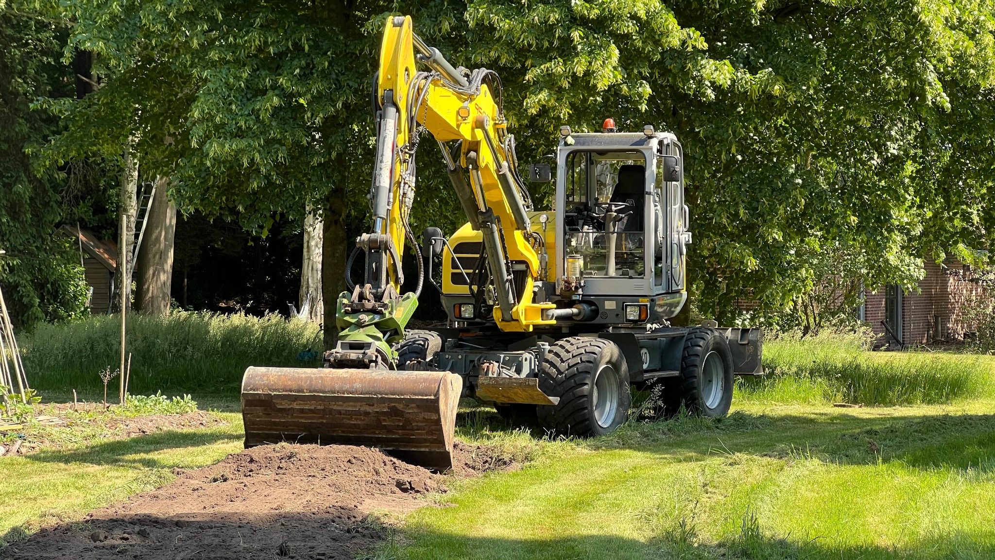 Żółto-szara koparka kołowa na trawiastym terenie z wykopanym wąskim rowem. W tle drzewa i budynek z cegły. Słoneczny dzień, widok z bliska.